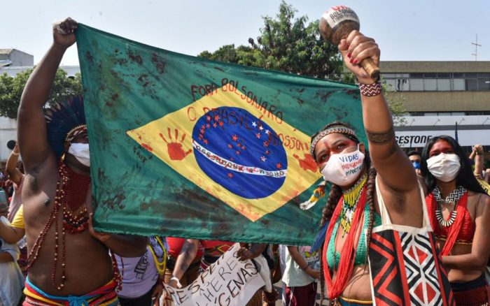 Indígenas seguram Bandeira do Brasil manchada de vermelho, imitando sangue, com frase 