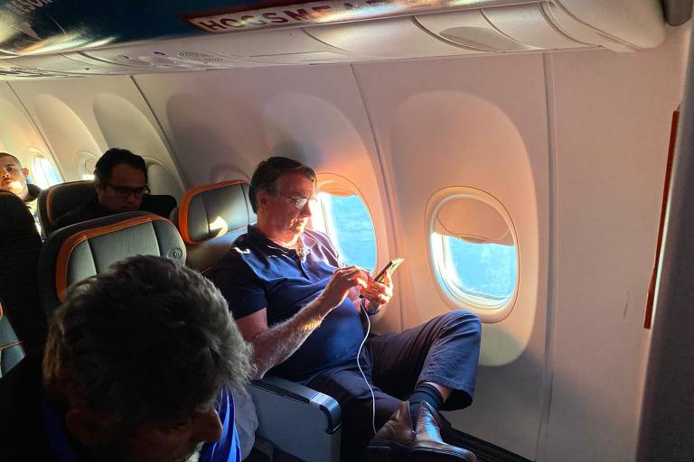 O ex-presidente Jair Bolsonaro usa celular em avião no qual volta ao Brasil. (Anna Virginia Balloussier/ Folhapress)